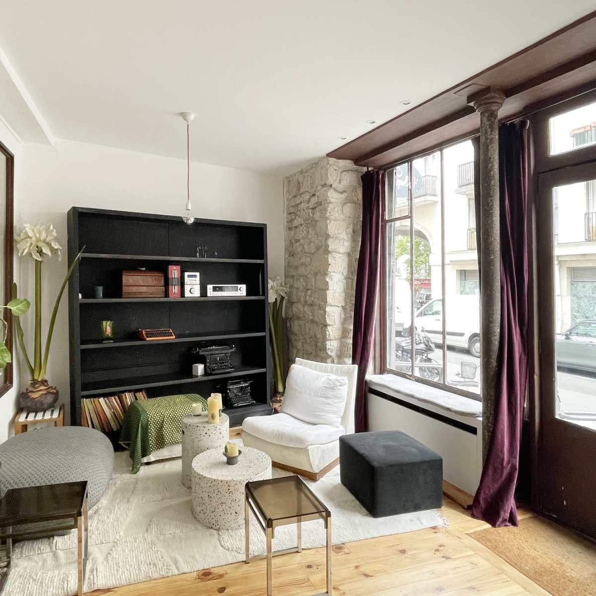 A cozy living room with a black bookshelf, minimalist furniture, a coffee table, and large windows allowing natural light to enter.