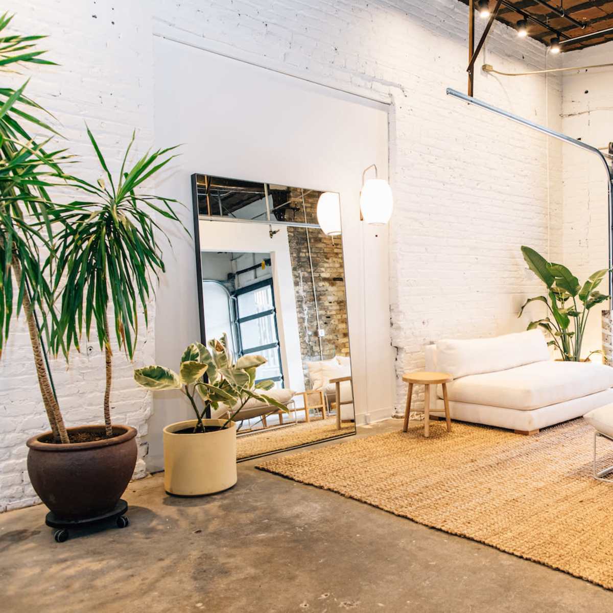 A spacious living area with a white couch, a mirror, and various plants set against a backdrop of exposed brick and concrete floors, with large garage doors allowing natural light.