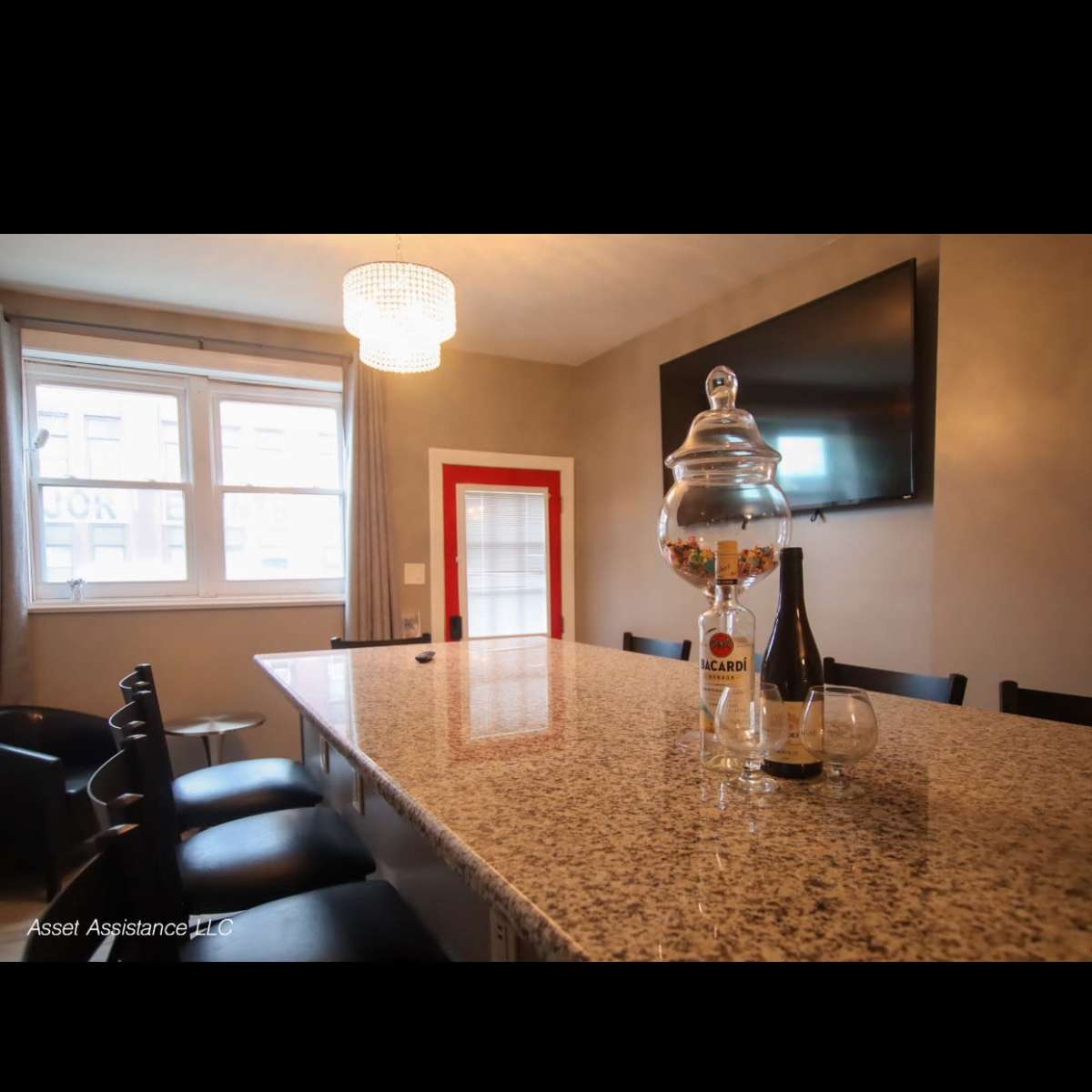 A long granite countertop with a beverage dispenser and a bottle is set against a wall featuring a large TV and a red door.