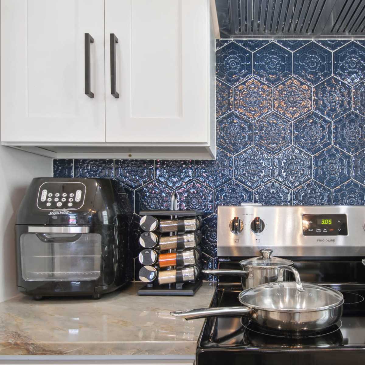 A modern kitchen with a stainless steel stove, pots on the burner, an air fryer on the counter, and a spice rack beside a blue tiled backsplash.