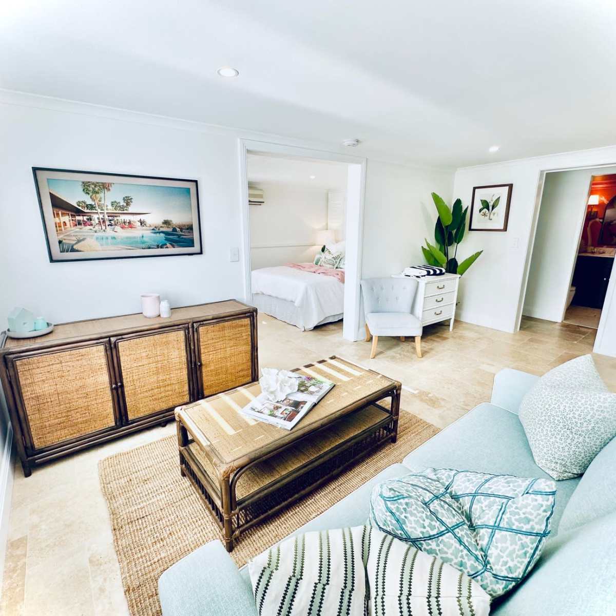 A bright and modern living space featuring a light-colored sofa, a wicker coffee table, and an open layout that connects to a bedroom area.
