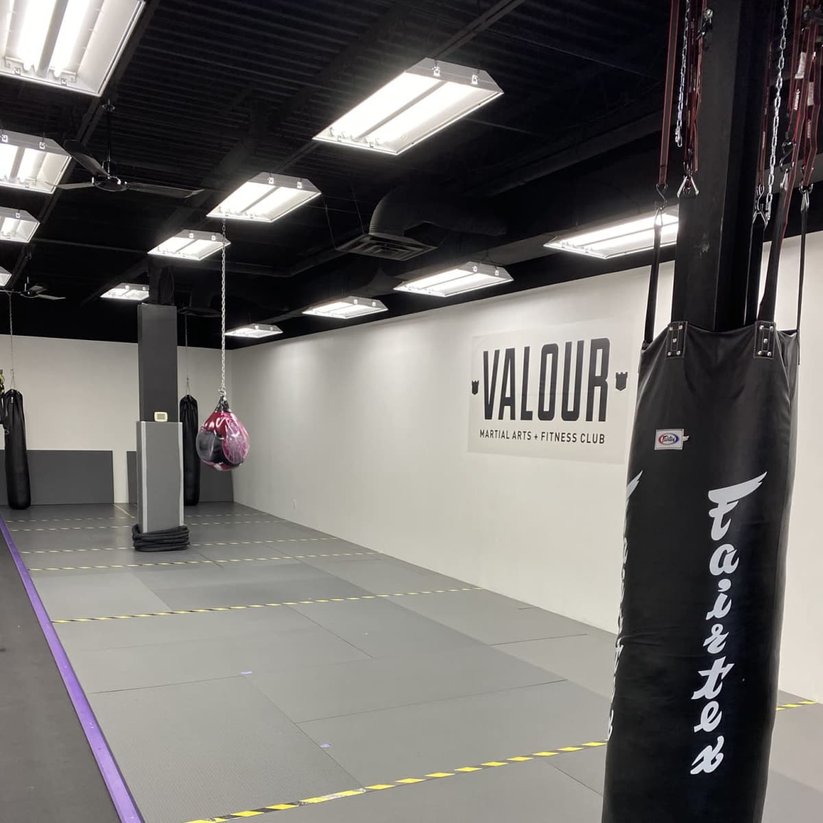 An empty martial arts gym with padded flooring, a punching bag, and a wall displaying the word "VALOUR."