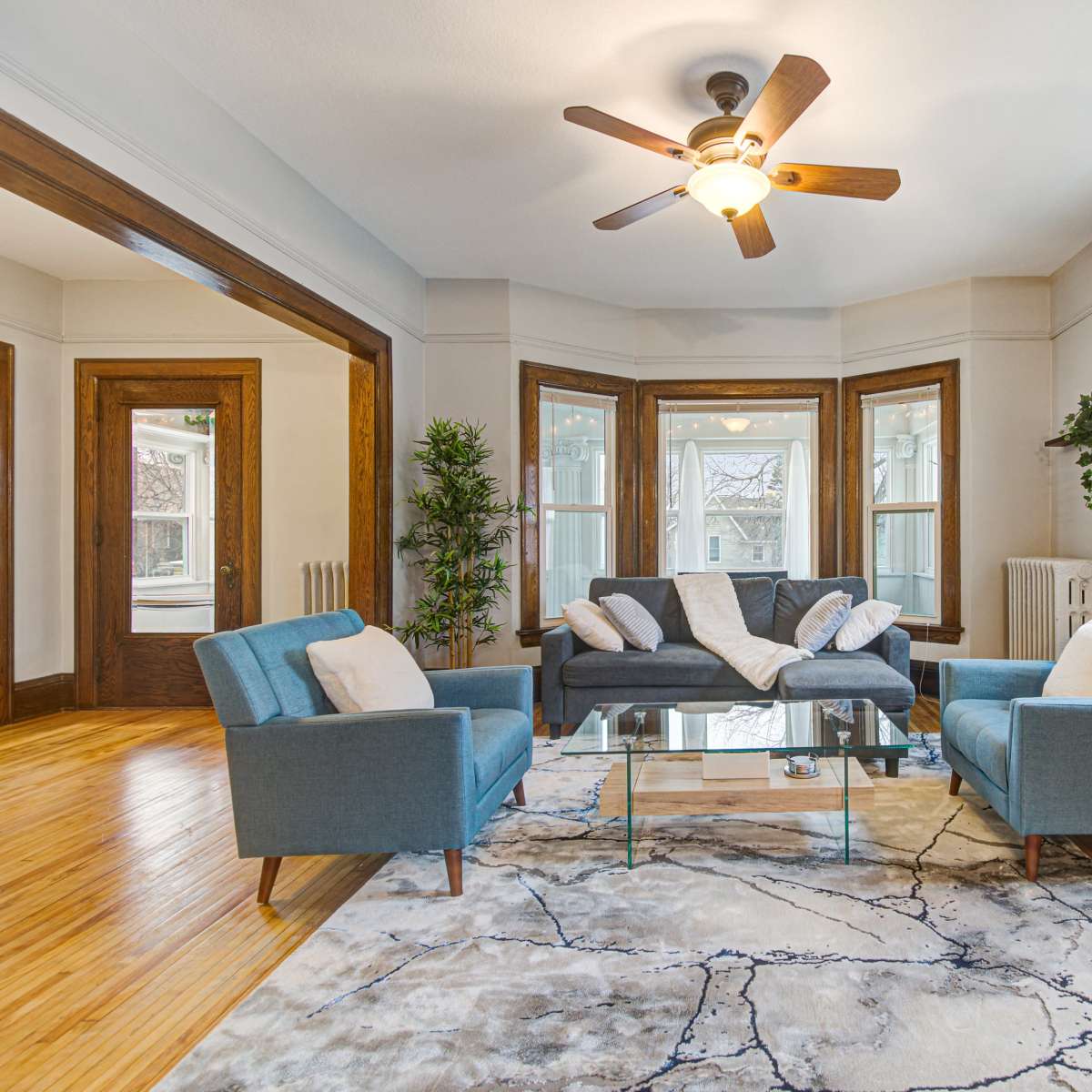 A living room with a light-colored rug, a glass coffee table, two blue armchairs, and a gray sofa, all arranged around a ceiling fan and bright windows.