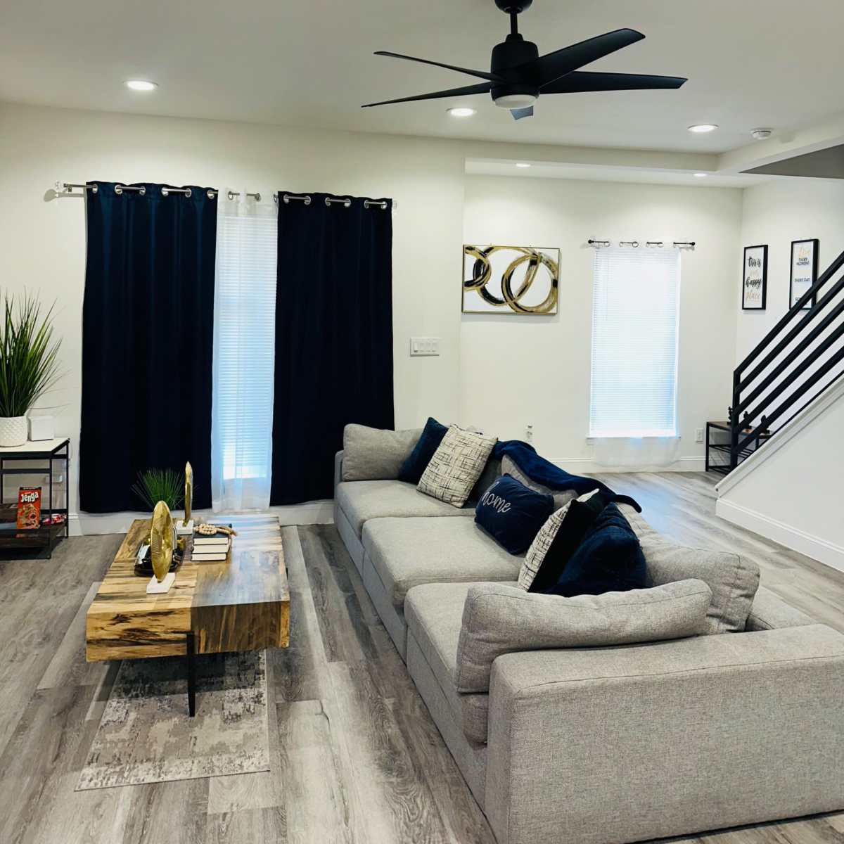 A modern living room features a gray sectional sofa, a wooden coffee table, dark curtains, and a ceiling fan, with stairs and wall art in the background.