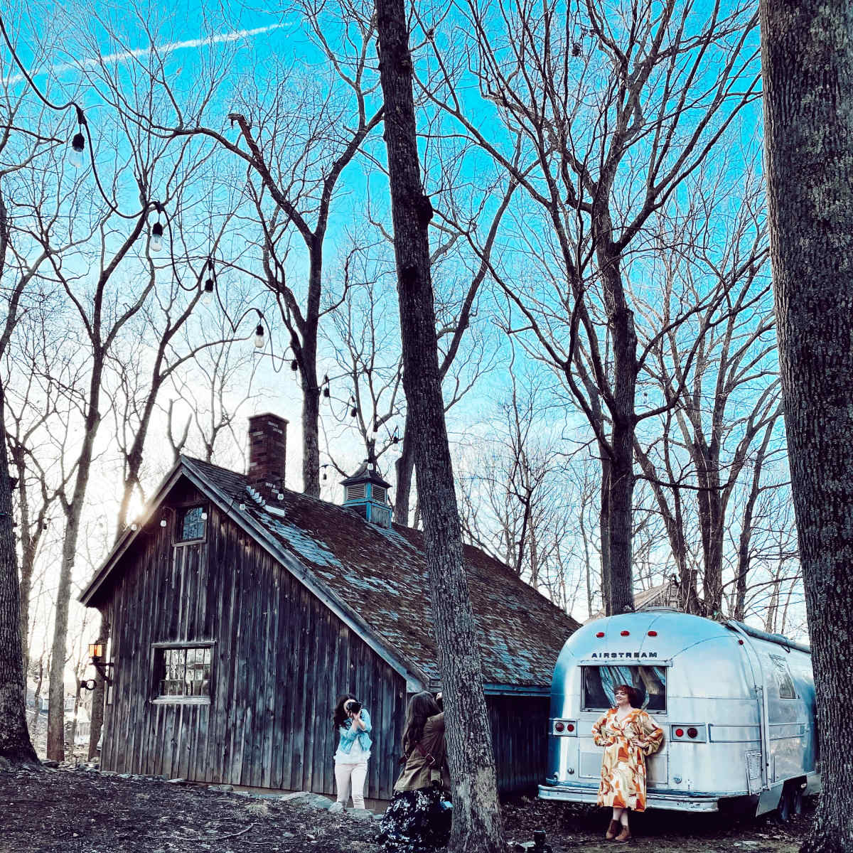 A silver Airstream trailer is parked next to a rustic wooden cabin surrounded by bare trees.