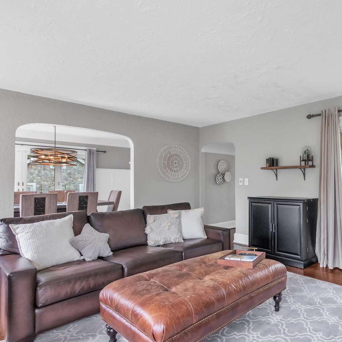 The living room features a brown leather sofa with decorative pillows, a wooden ottoman, and a large window offering a view of the outdoors.