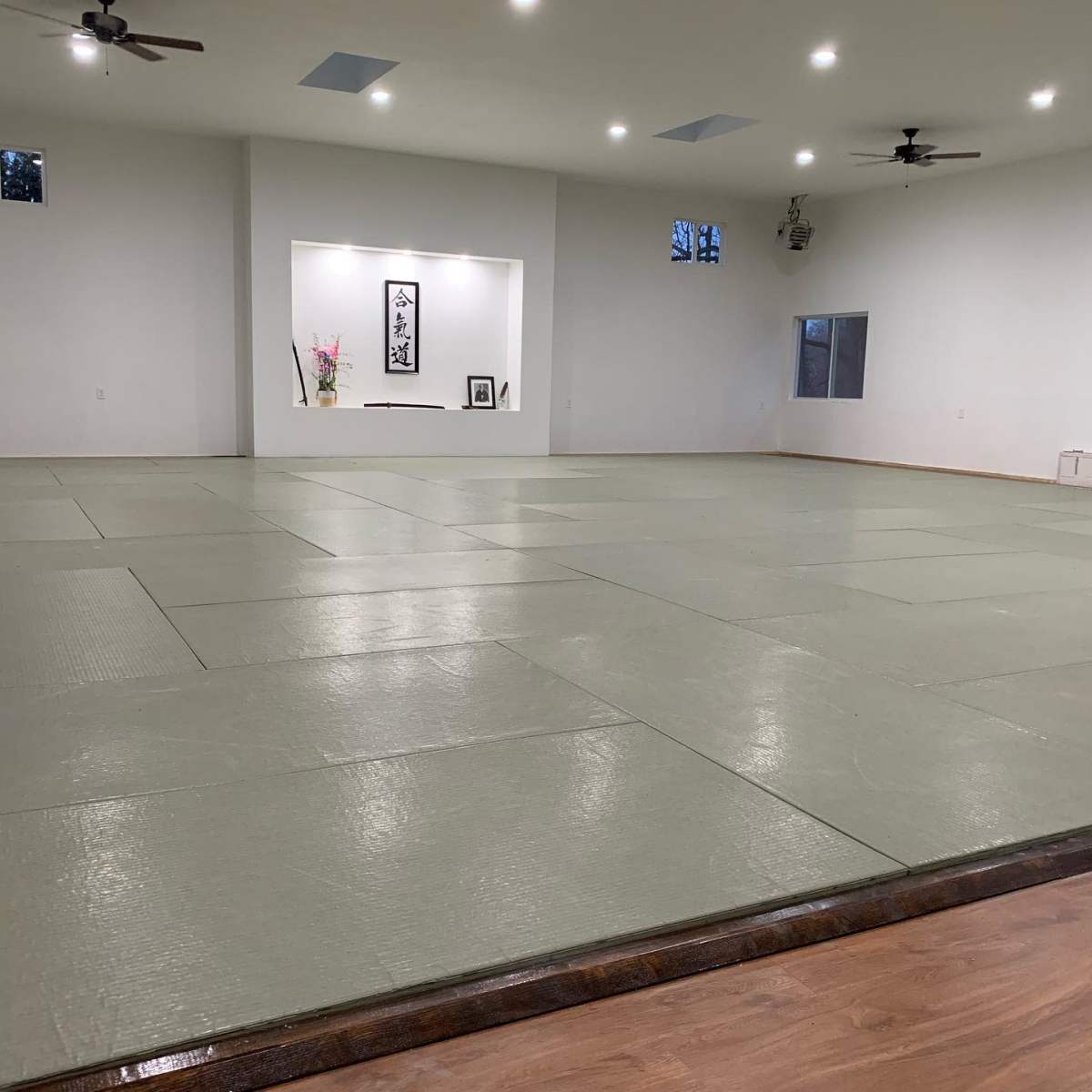 A spacious, empty room with green tatami mats and a decorative wall featuring a framed art piece and a vase with flowers.