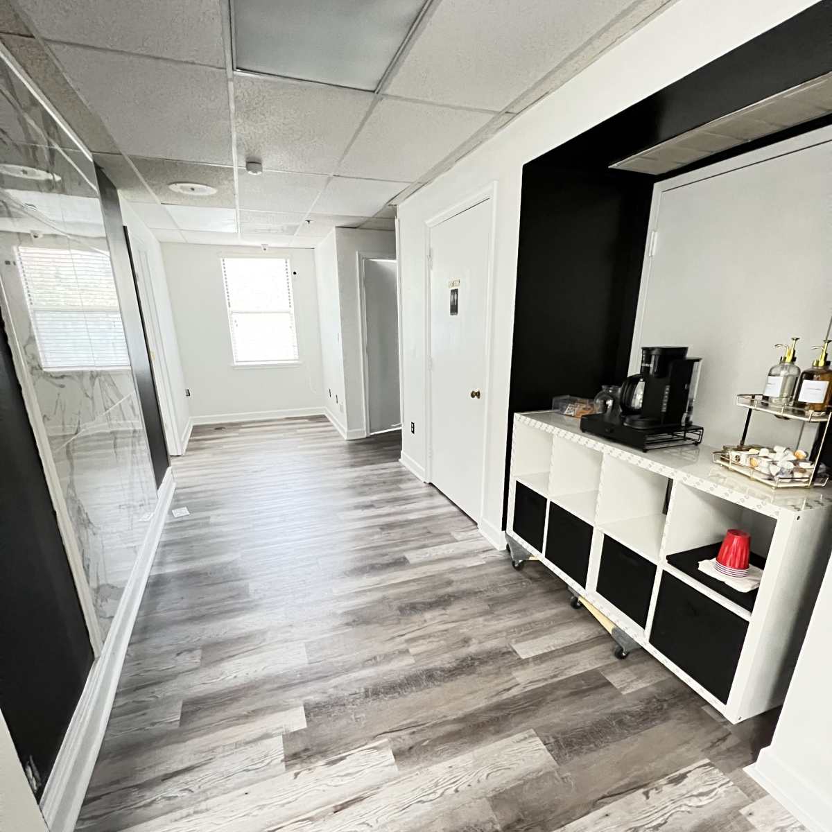 A hallway with light-colored wooden flooring, leading to a well-lit room at the far end, featuring a coffee station on the right.