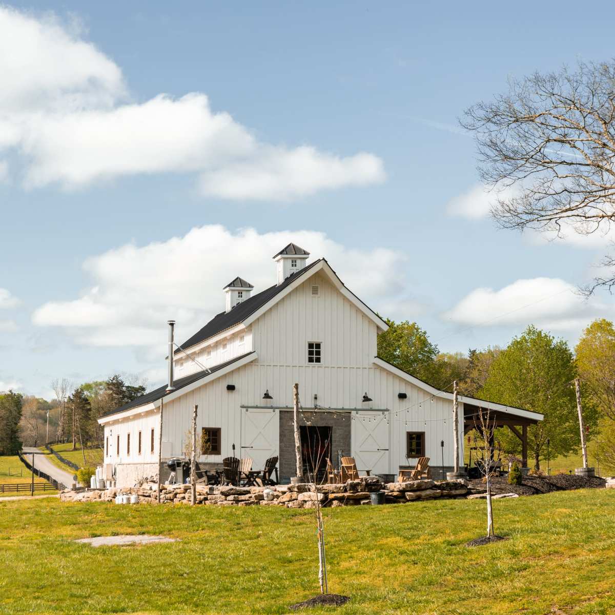 A white barn with a stone patio and nearby trees is set against a cloudy sky in a rural landscape.