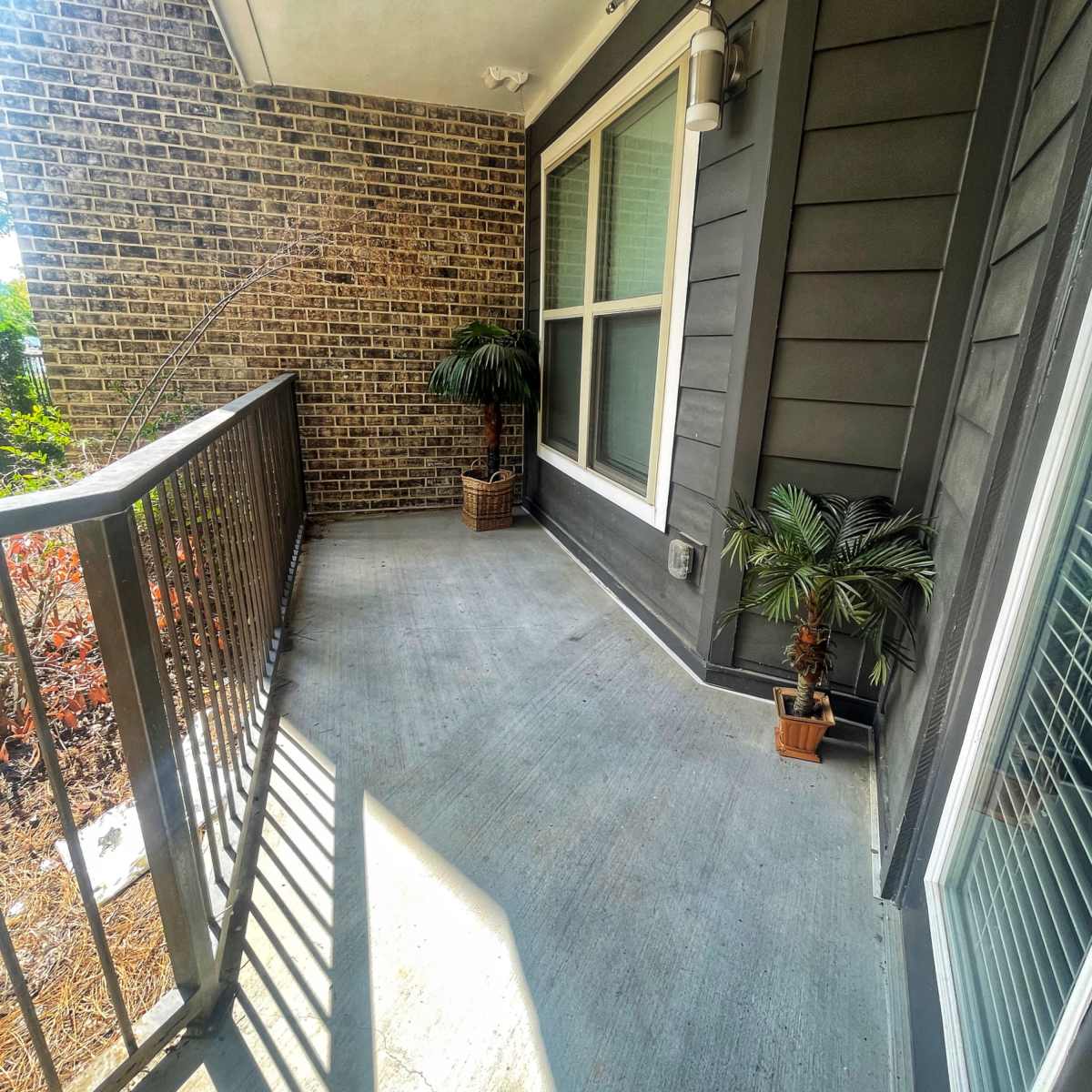A small covered balcony with potted plants and a railing, adjacent to a brick wall.
