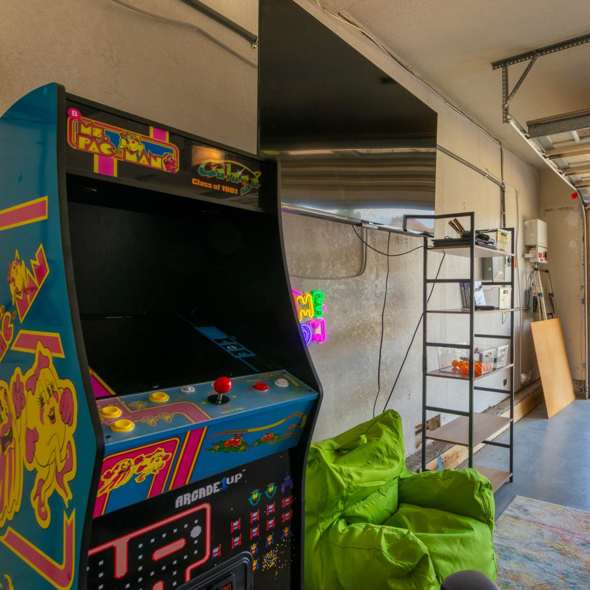 A garage is equipped with an arcade machine, gaming chair, and vibrant bean bags, with a view of a residential street outside.