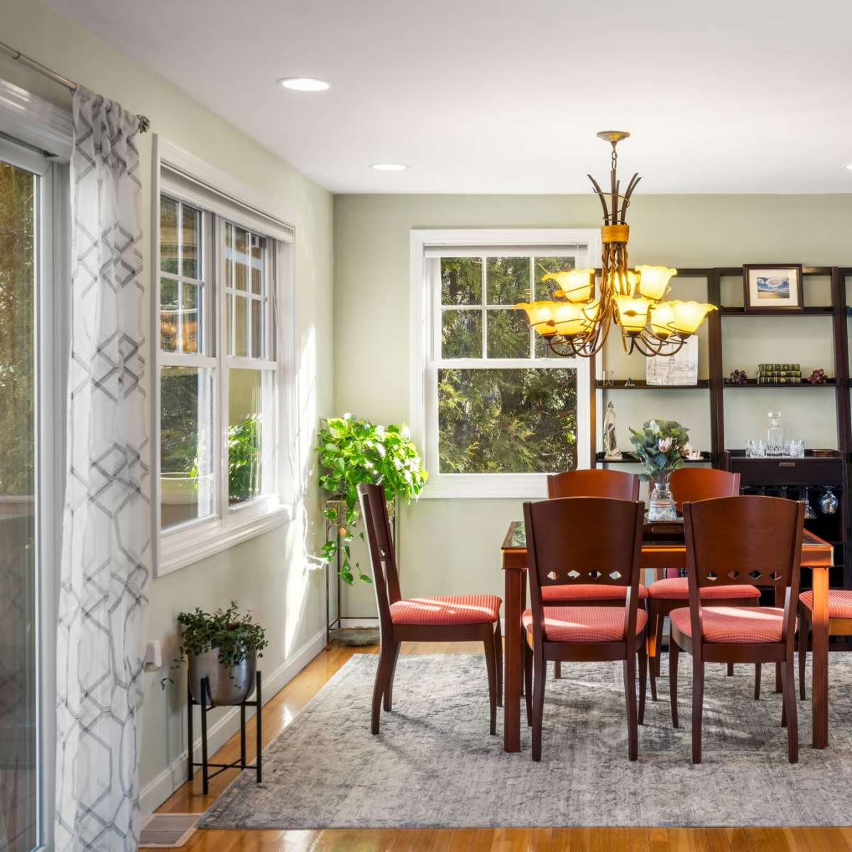 A dining area features a wooden table surrounded by four chairs, illuminated by a chandelier, with large windows and a shelf displaying decorative items.