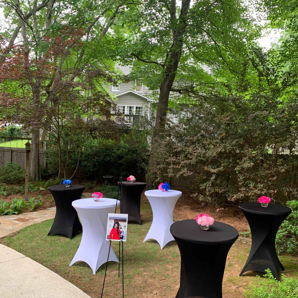 The scene shows several round tables with fitted tablecloths in a garden, each adorned with floral arrangements, surrounded by trees and shrubs.