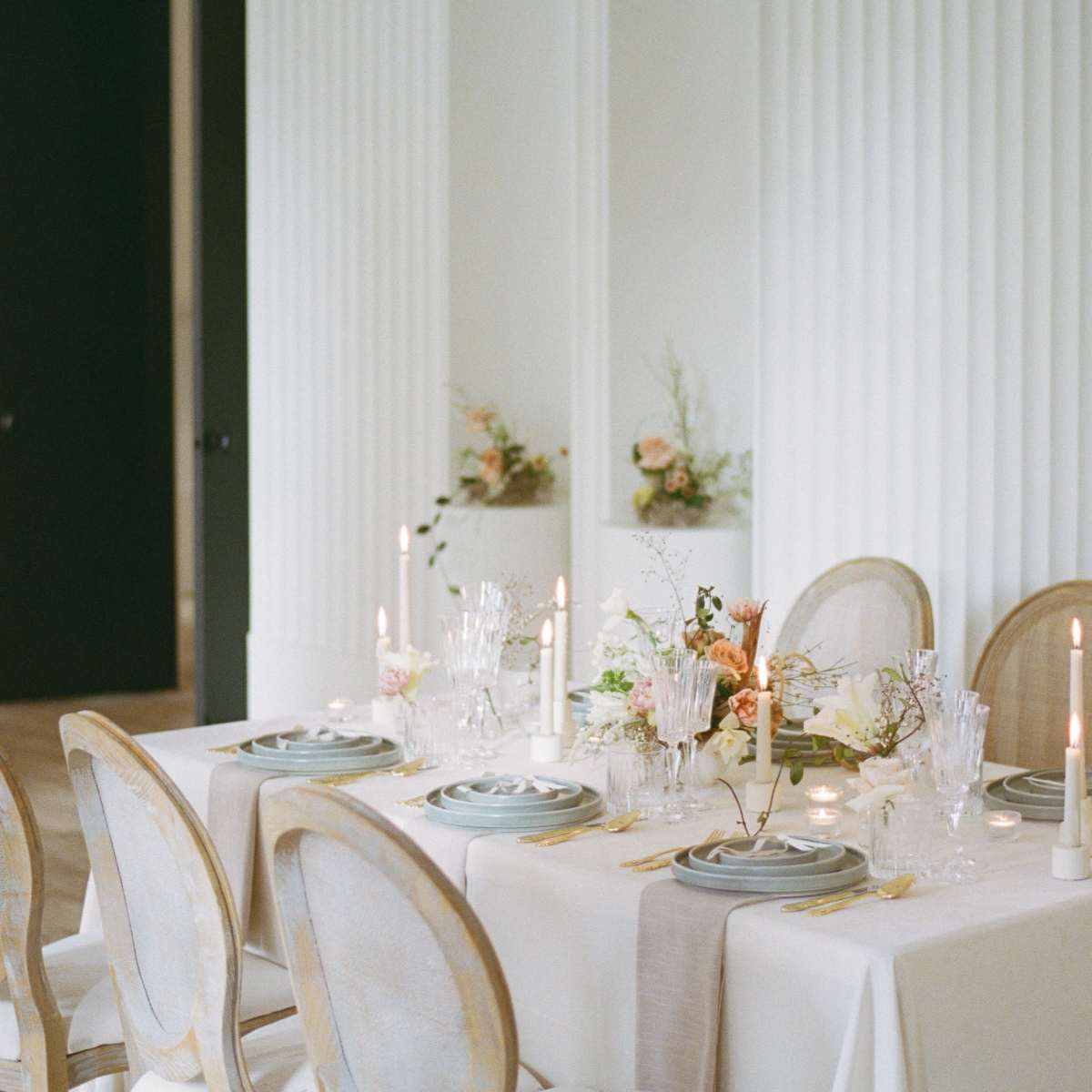 A dining table is elegantly set with candles and floral arrangements against a backdrop of textured white walls.
