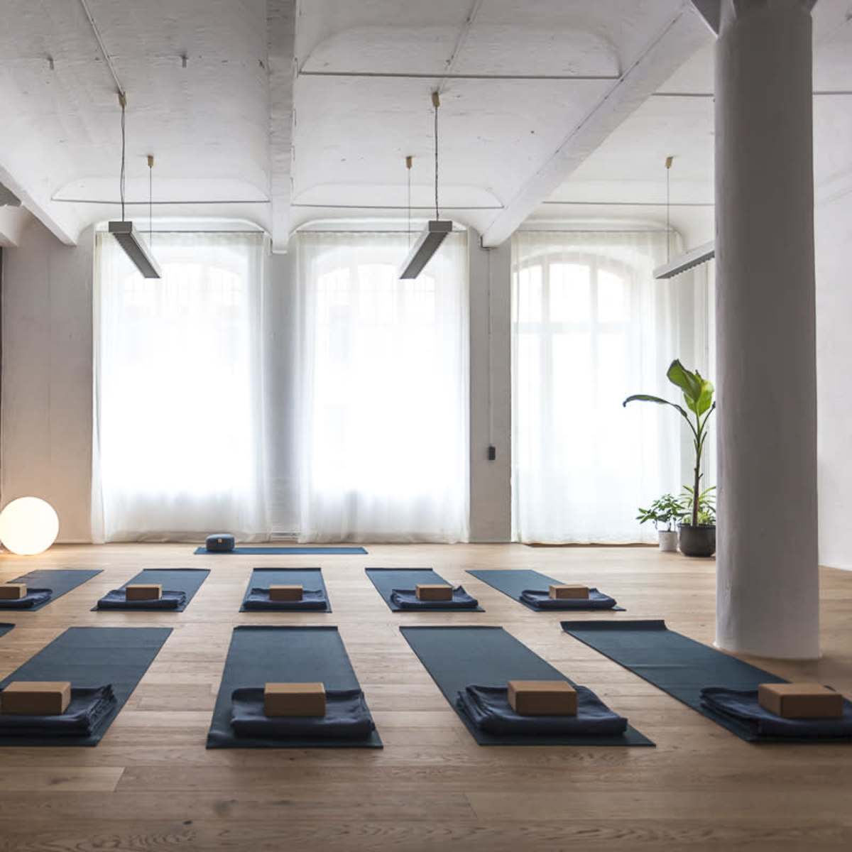 A yoga studio with neatly arranged mats and blocks on a wooden floor, illuminated by natural light filtering through large windows.