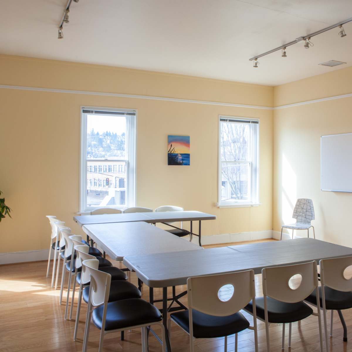 A well-lit meeting room features a long table surrounded by chairs, with windows providing natural light and a whiteboard on one wall.