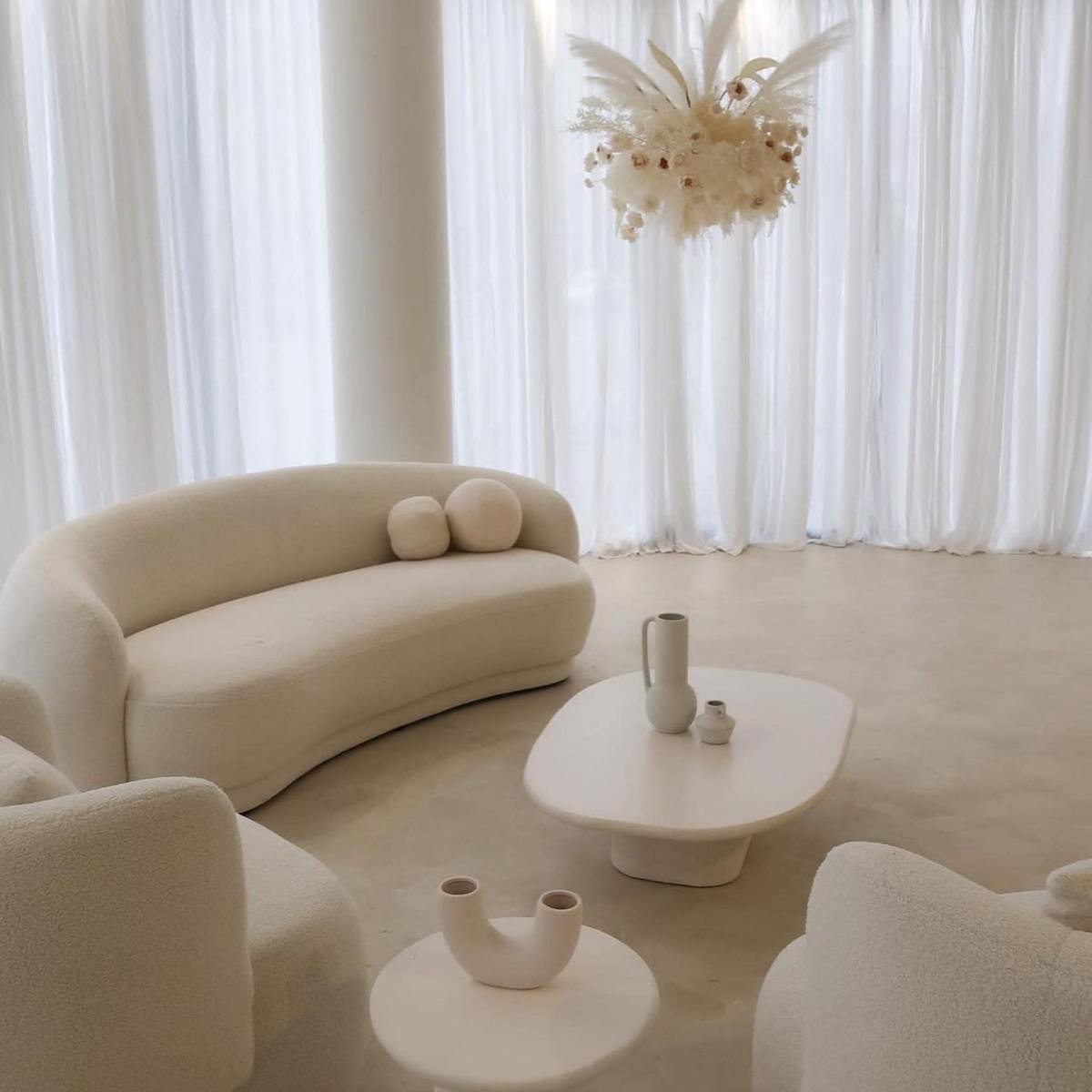 A minimalist living space with a curved white sofa, a round coffee table, and a ceiling-mounted floral arrangement, all complemented by sheer white curtains.