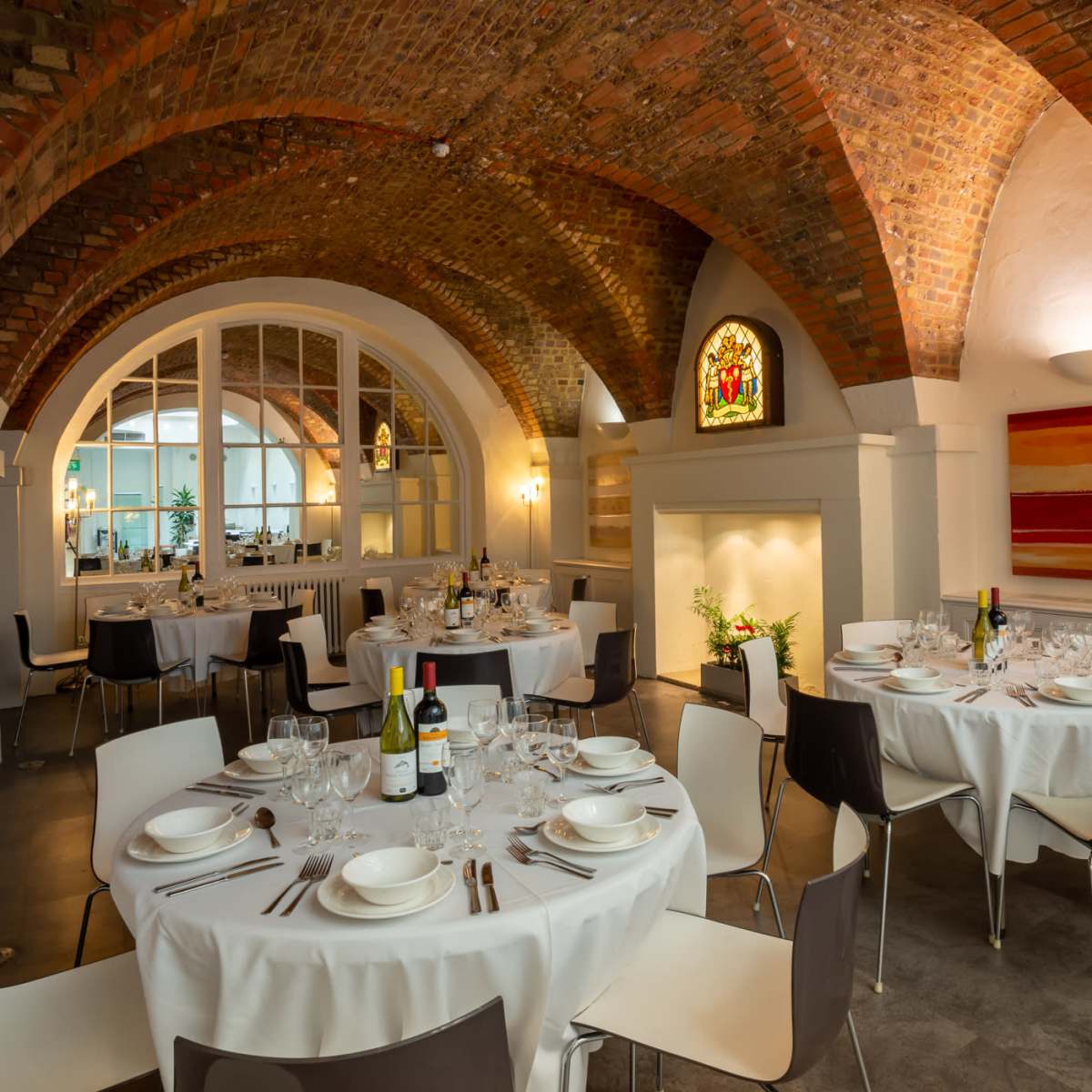 A dining area in a restaurant with arched brick ceilings, round tables set with white tablecloths, and large windows letting in light.
