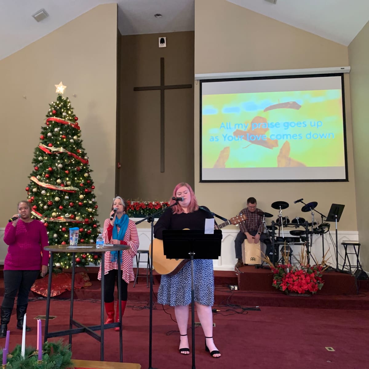 A group of musicians performs in front of a decorated Christmas tree in a church setting, with a projected screen displaying lyrics in the background.
