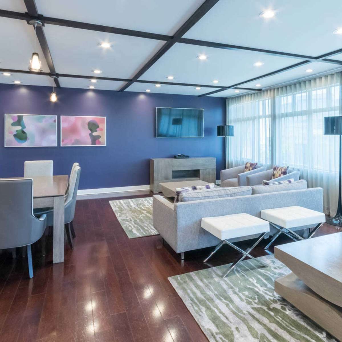 A modern living room features a dining area, comfortable seating, and large windows with sheer curtains, set against a purple accent wall.
