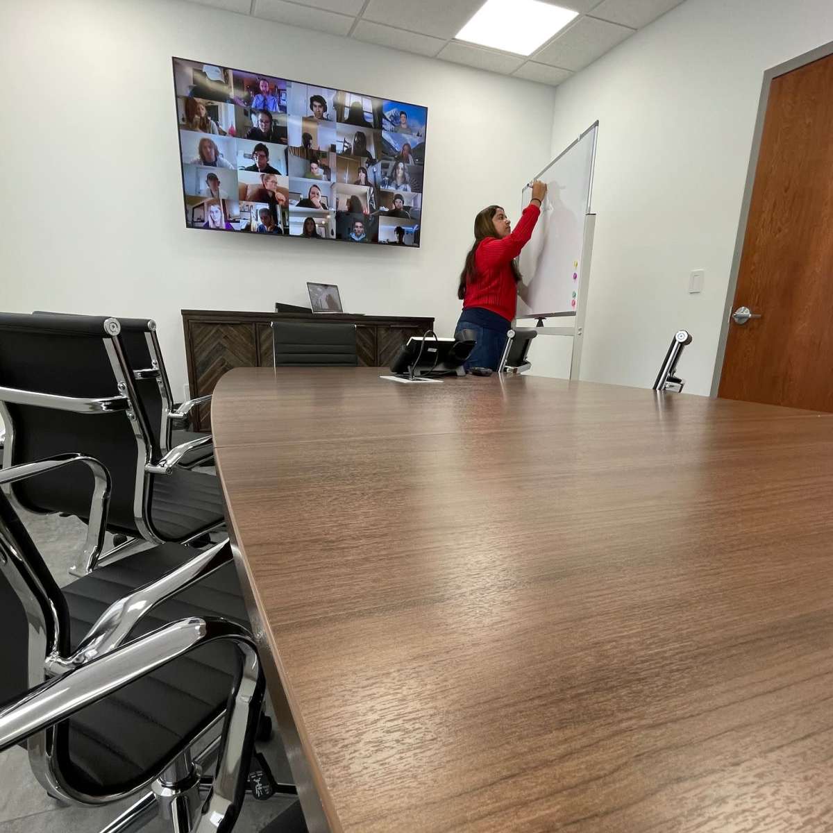 A person writes on a whiteboard in a conference room equipped with a large screen displaying a video call with multiple participants.