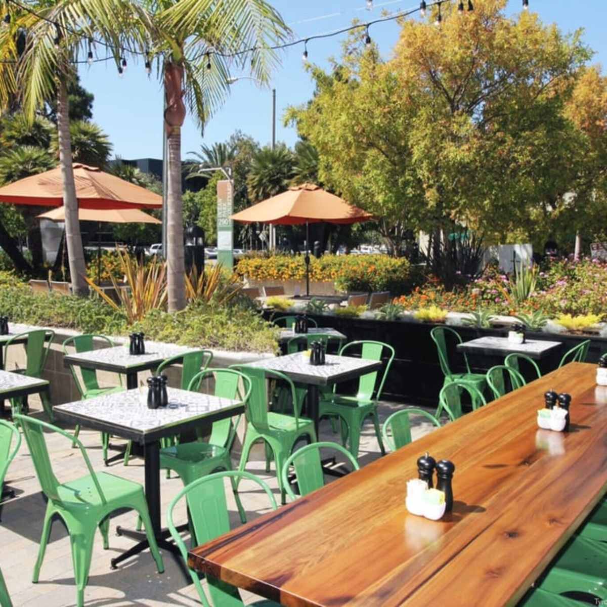 A patio area features green metal chairs and wooden tables set up under orange umbrellas surrounded by plants and flowers.