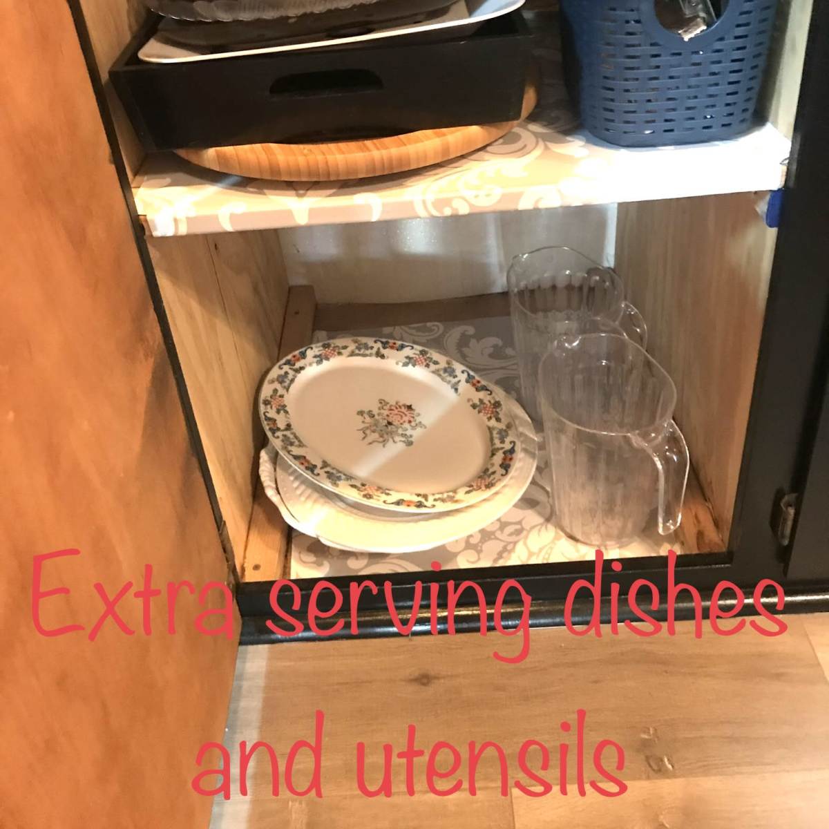A wooden cabinet shelf holds a variety of serving dishes, utensils, and glass pitchers.