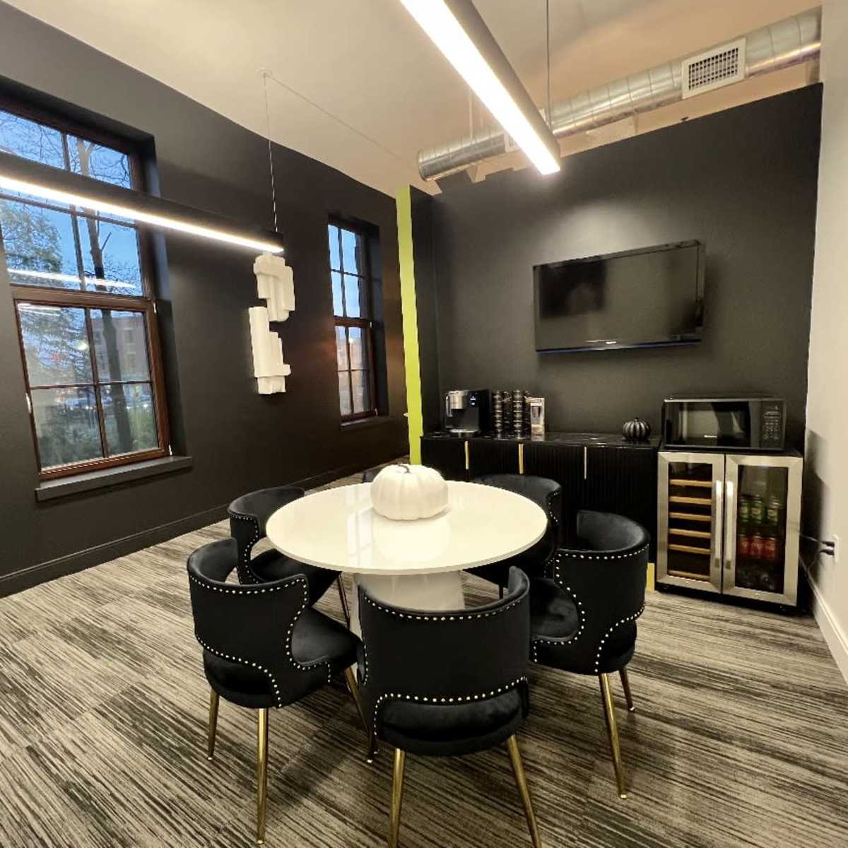 A modern meeting room featuring a round white table surrounded by black chairs, a wall-mounted TV, and a kitchenette area with a microwave and beverage cooler.