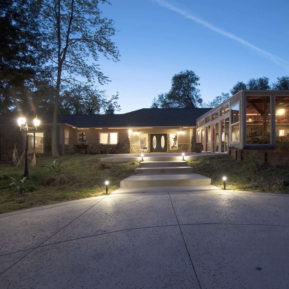 A well-lit pathway leads to a modern home surrounded by trees, with a glass-enclosed patio visible on one side.