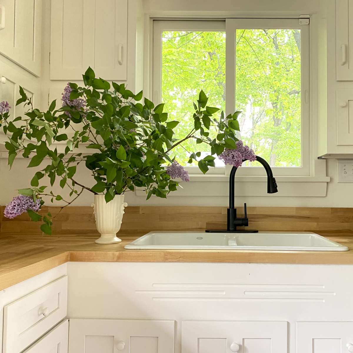 A kitchen features a window with greenery outside and a vase of lilacs on the countertop near a black sink.