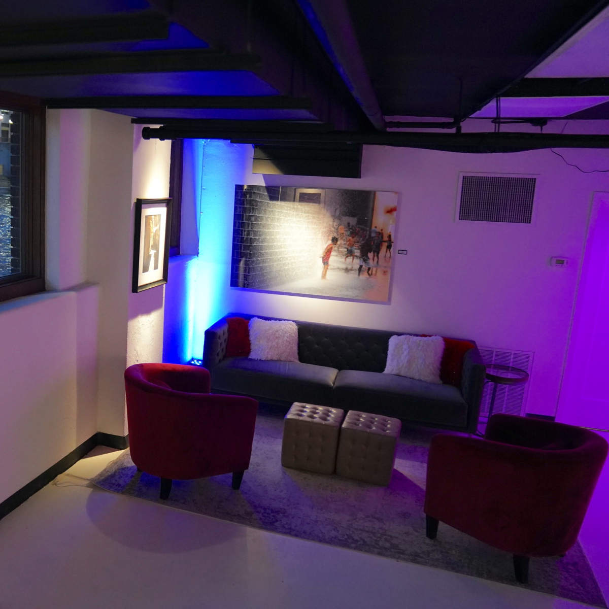 A modern lounge area with a dark sofa, two red chairs, and blue ambient lighting beside large windows overlooking a body of water.