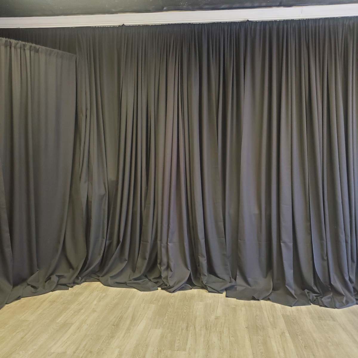 A corner of a room with floor-to-ceiling black curtains draped along the walls, contrasting with a wooden floor.