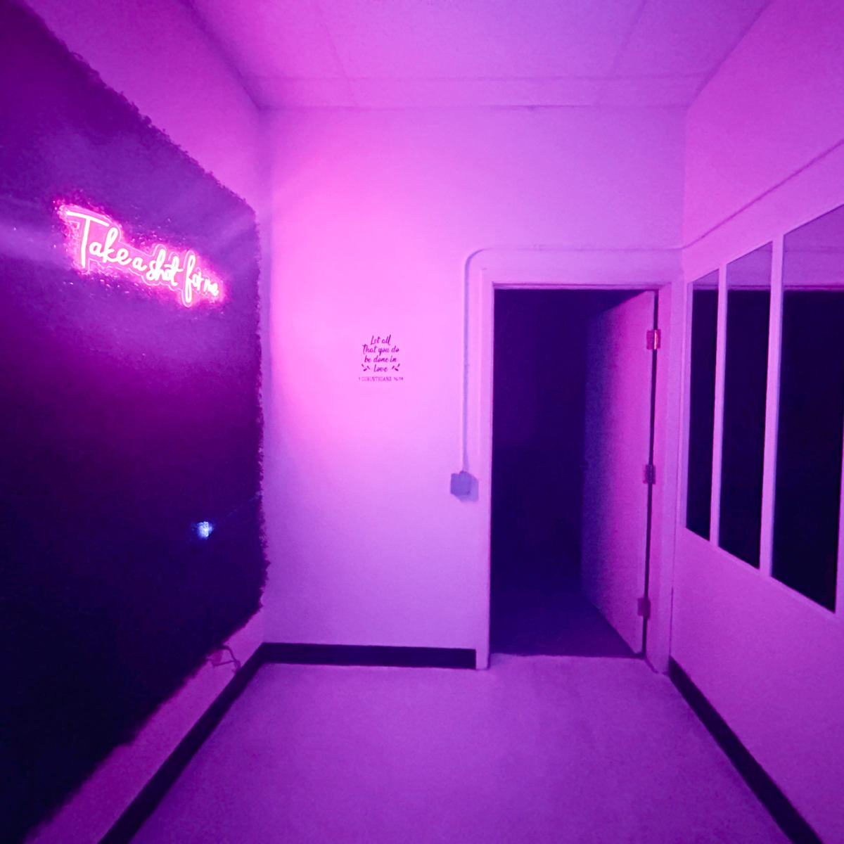 A dimly lit room features a neon sign reading "Take it in" on one wall, with a mirrored surface opposite and an open doorway leading to a dark area.