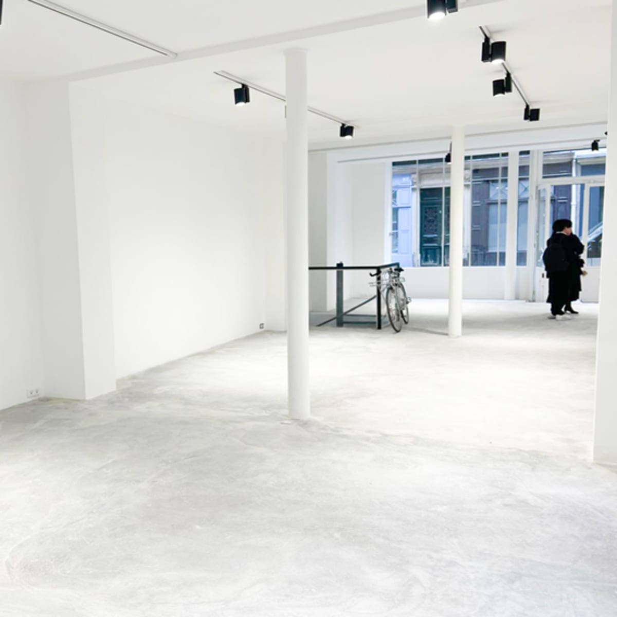 A spacious, minimally furnished room with white walls, concrete flooring, and a large window, where a person in dark clothing stands near a bike.