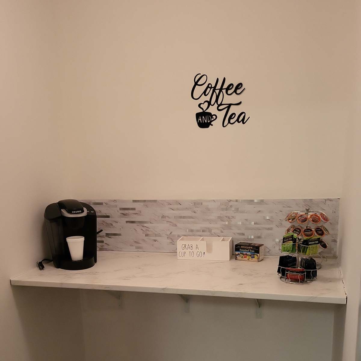 A small nook features a coffee maker and a selection of snacks against a white wall with the words "Coffee and Tea" above a marble-inspired countertop.