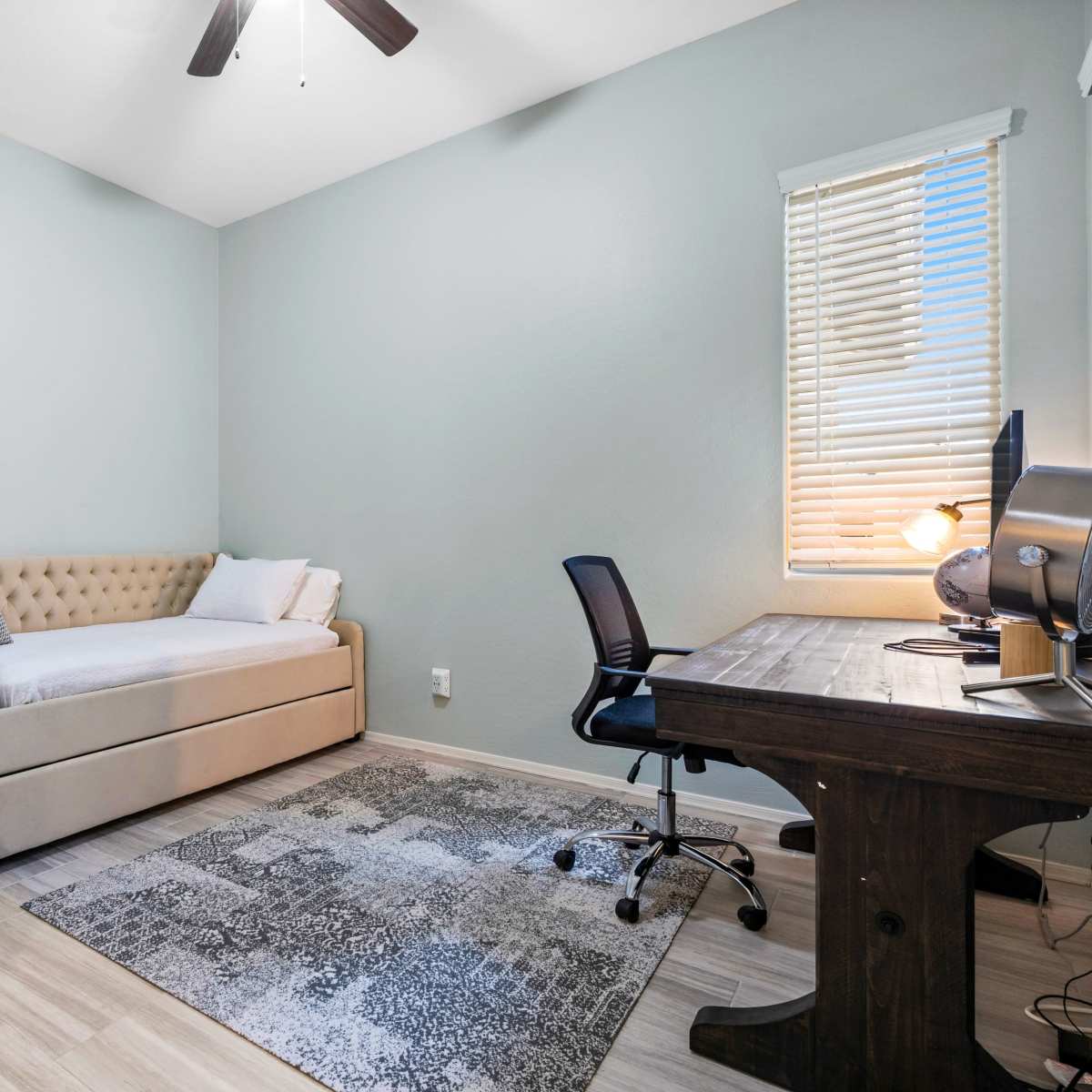 A small bedroom with a daybed, a desk with a computer chair, and a decorative cabinet against a light blue wall.