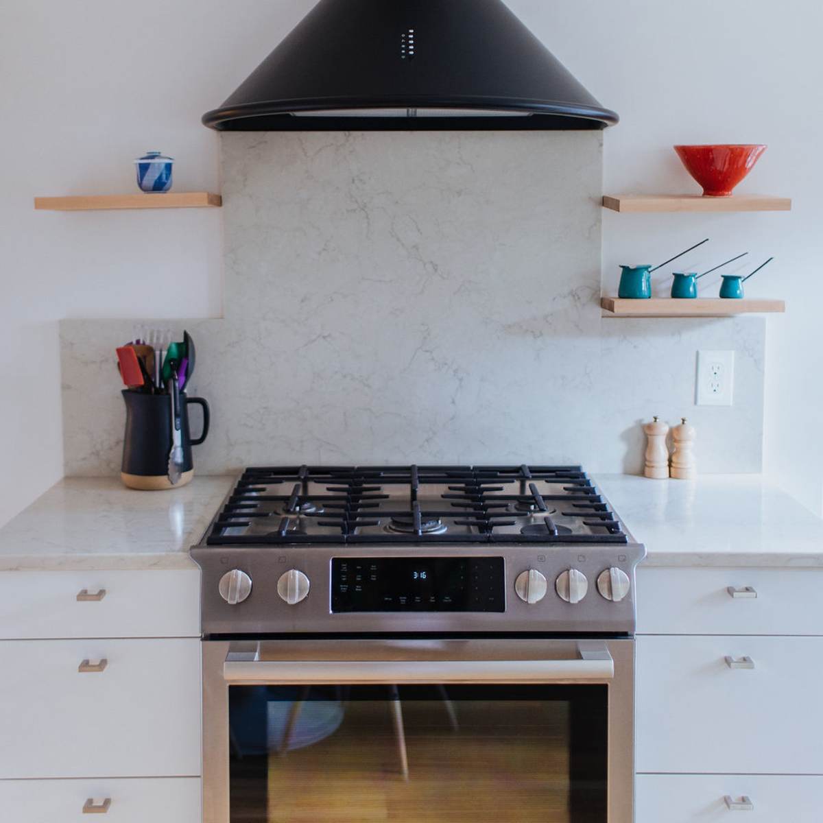 A modern kitchen with a stainless steel stove, a black hood, and open shelves displaying various kitchen utensils and colorful dishes.