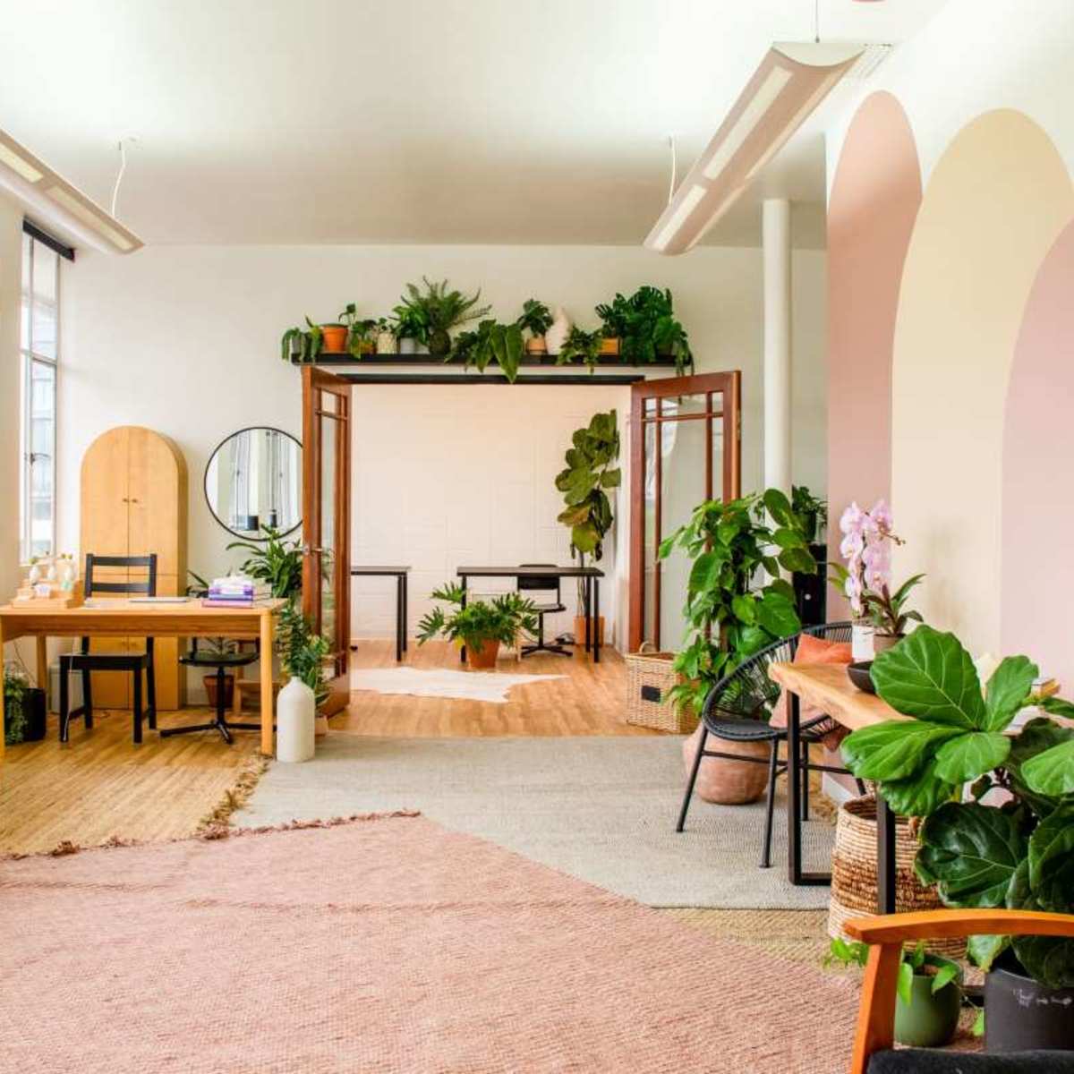 A bright, modern workspace with wooden furniture, plants, and decorative rugs arranged in an open layout.