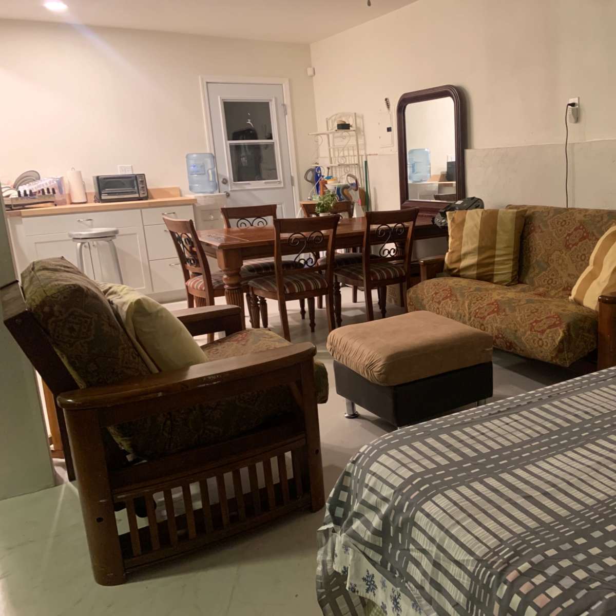 A living space with a small kitchen area, a dining table with six chairs, a sofa set, and a bed with a patterned cover.