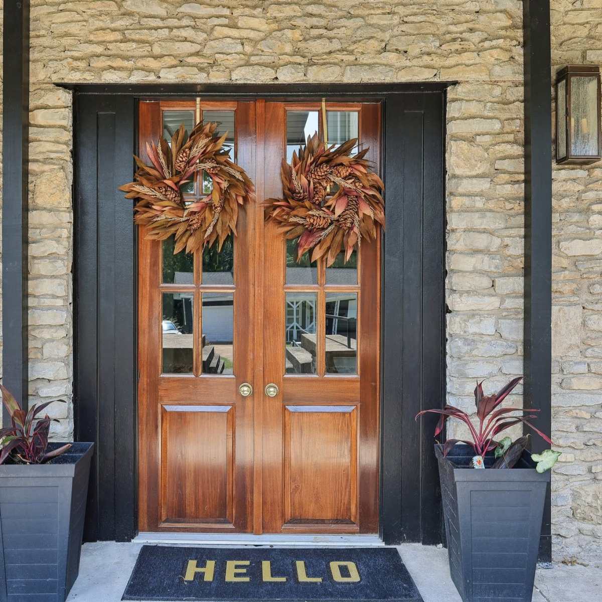 A pair of wooden double doors with glass panels, flanked by decorative wreaths, and surrounded by potted plants and a welcome mat.