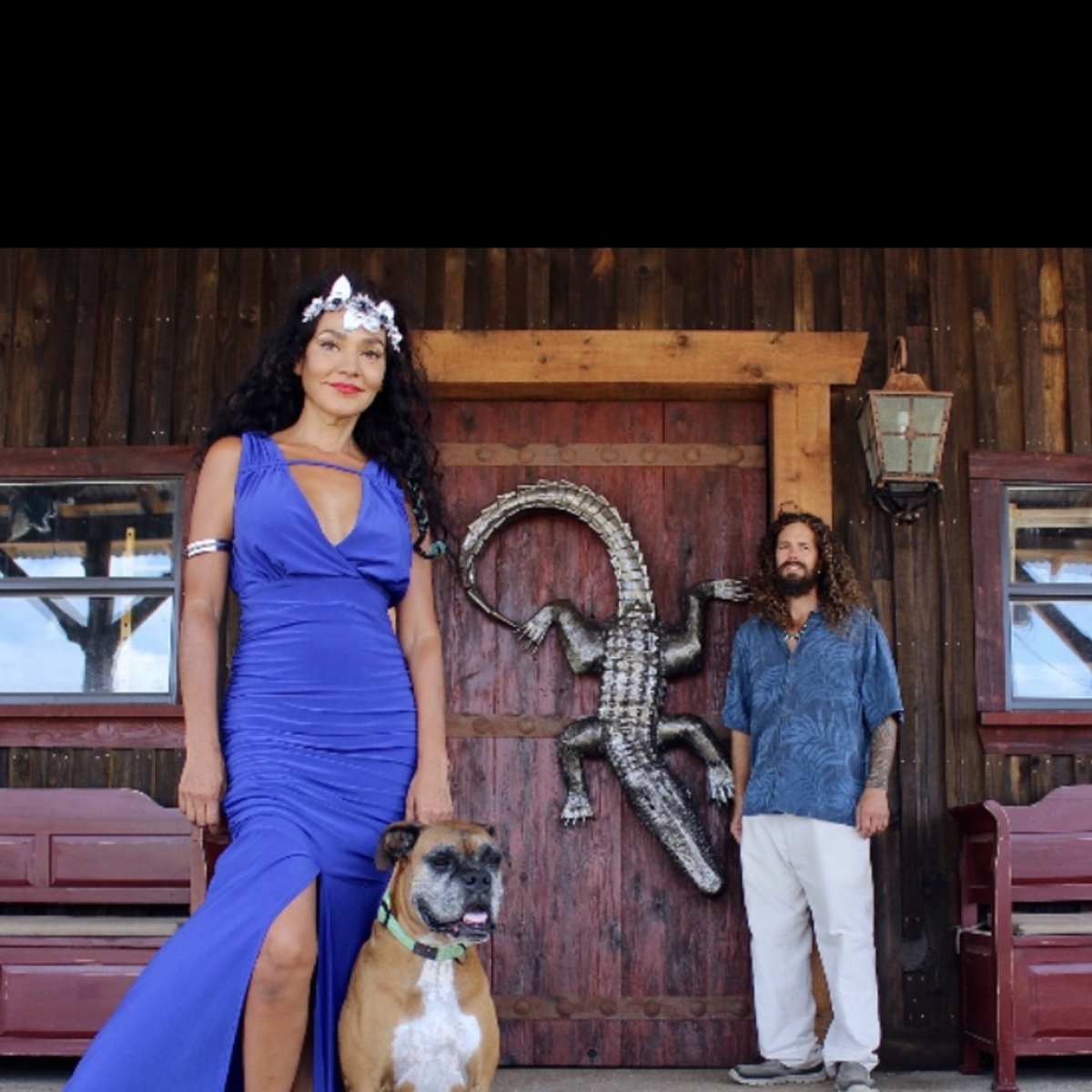 A woman in a blue dress stands next to a dog, while a man in a patterned shirt stands behind them in front of a wooden structure featuring a decorative alligator.