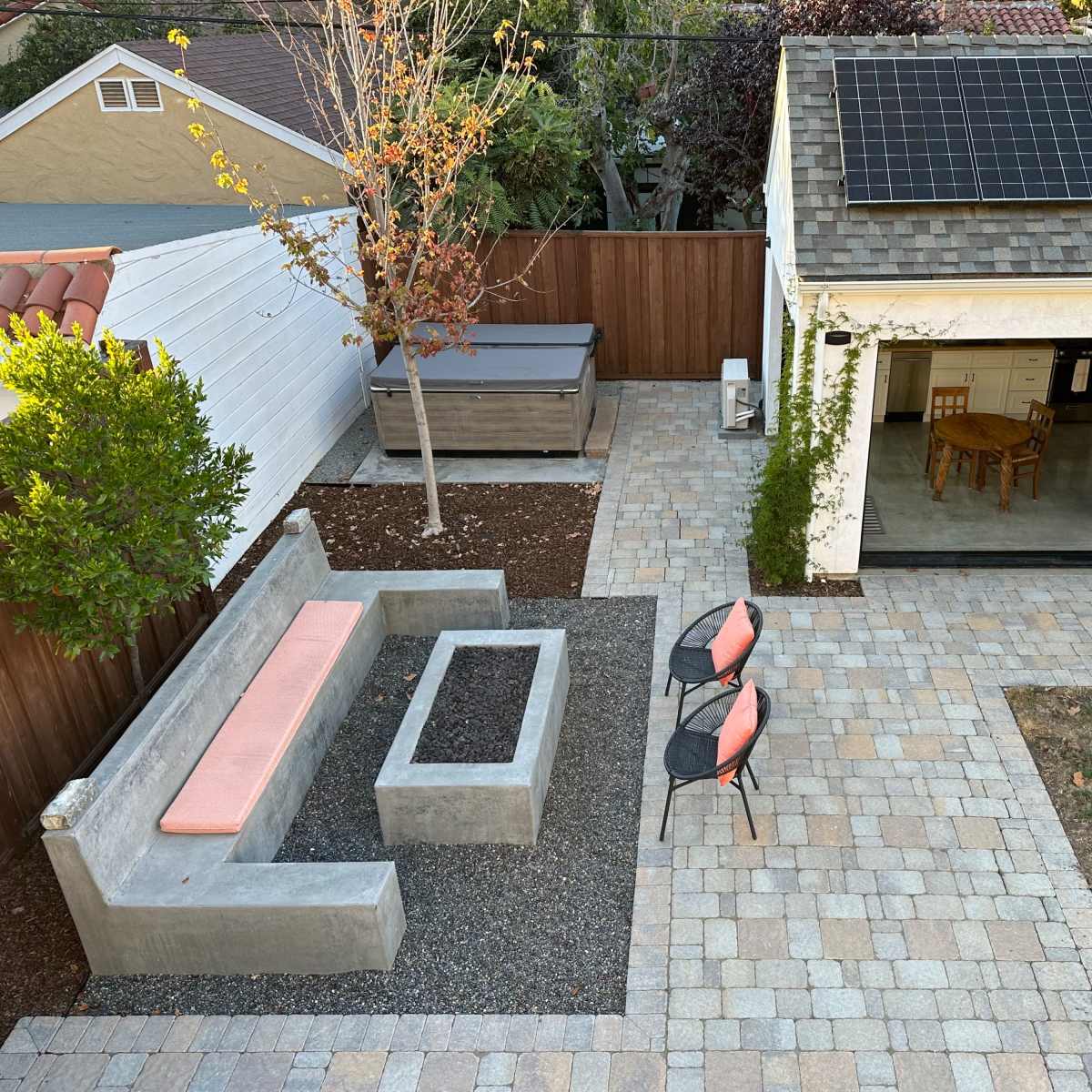 A landscaped backyard featuring a stone-paved patio with a concrete seating area, a fire pit, two chairs, and a building with a solar panel roof.