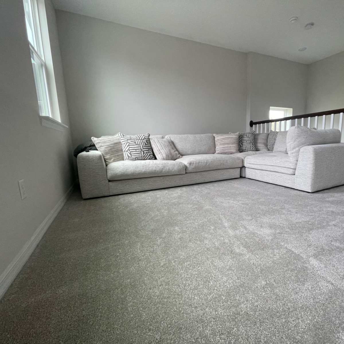 A large sectional sofa with decorative pillows is positioned in a carpeted room with light gray walls and a railing visible in the background.