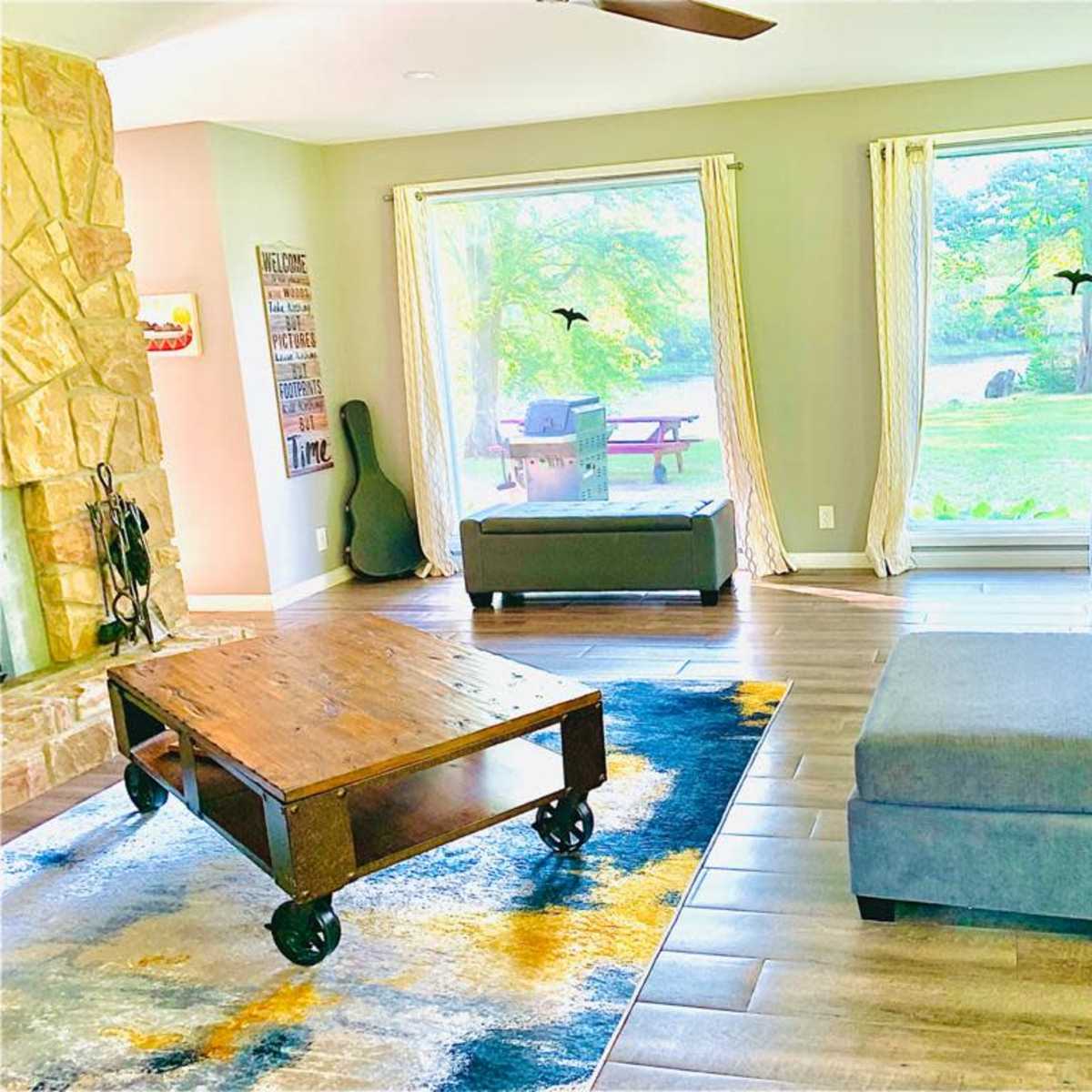 A living room features a stone fireplace, a large gray sectional sofa, a wooden coffee table on a patterned rug, and large windows overlooking a green yard with picnic tables.