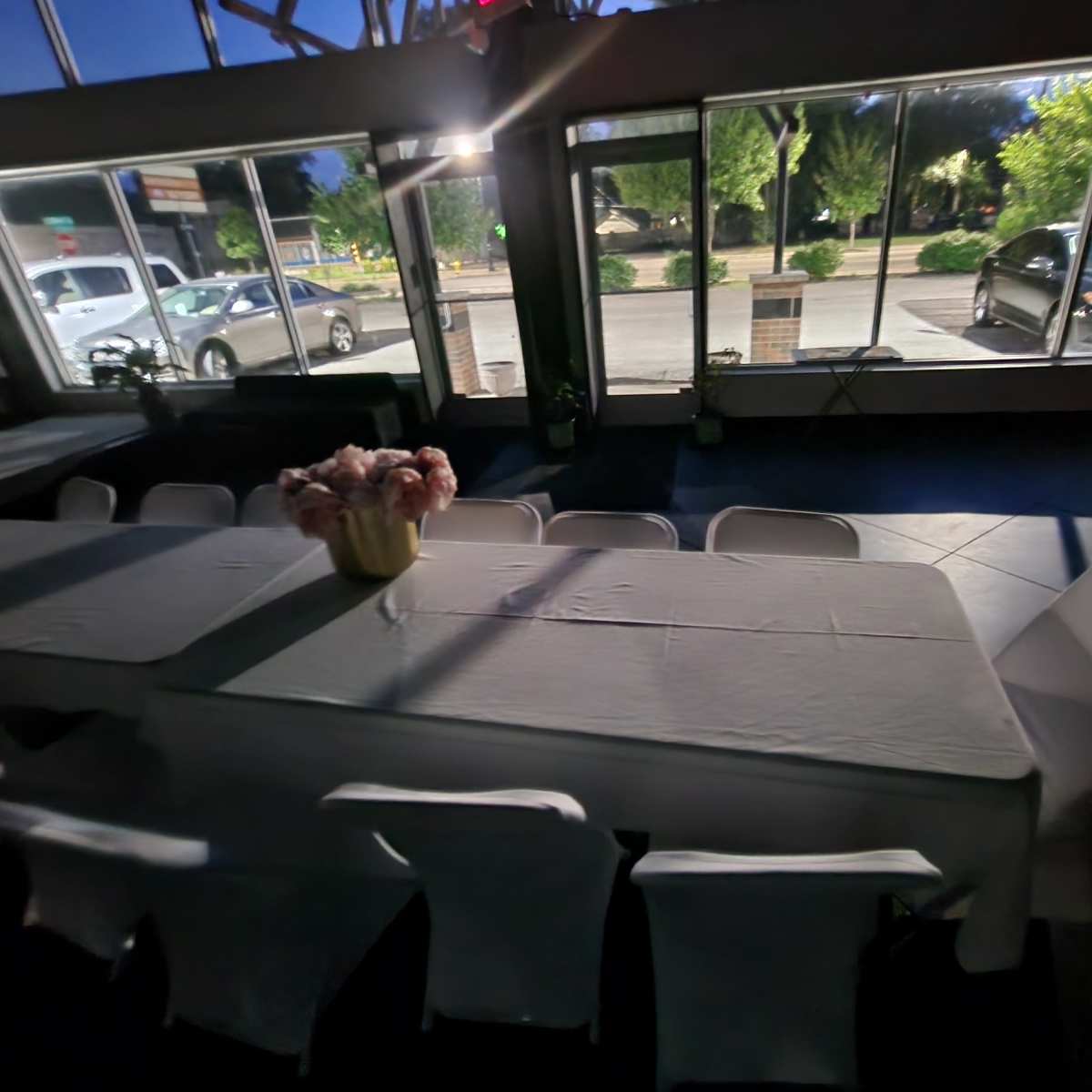 A long table covered with a white cloth is set in a room with large windows overlooking a parking lot and trees outside.