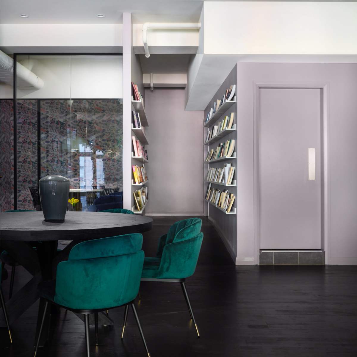 A modern interior features a round table with teal chairs and a wall-mounted bookshelf filled with books, alongside a door and a glass partition.