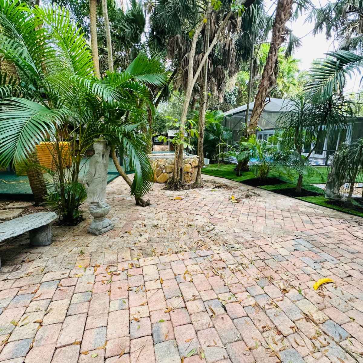 A landscaped garden area with brick pathways, stone benches, palm trees, and a small stone feature.