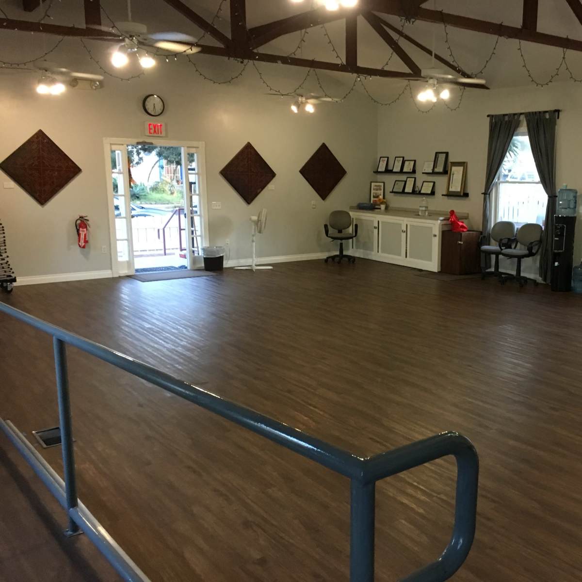 A spacious, empty room with a wooden floor, large windows, and an entrance, featuring a few chairs and a water cooler along one wall.