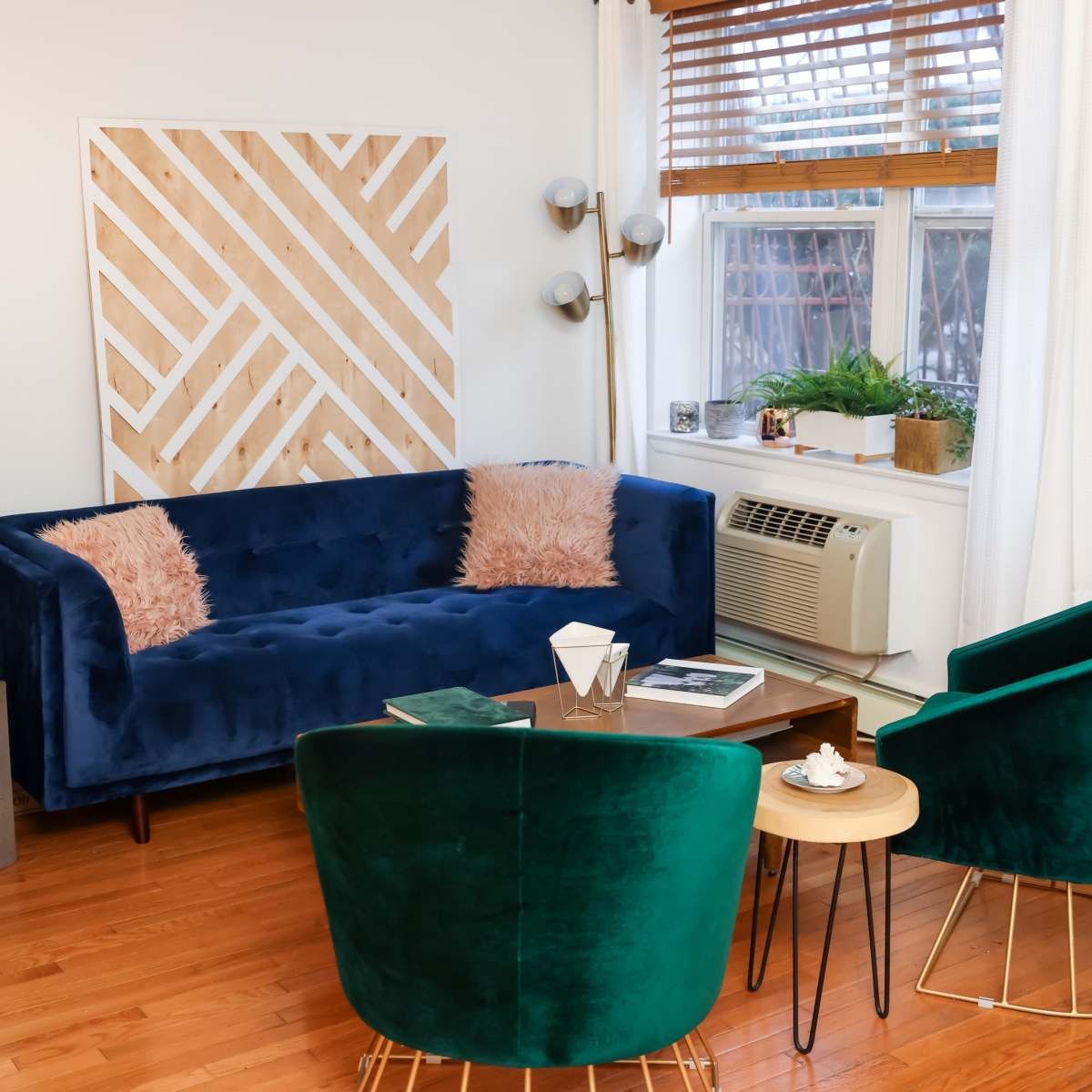 A modern living room with a blue velvet sofa, two green velvet chairs, a wooden coffee table, and a large window adorned with sheer curtains.