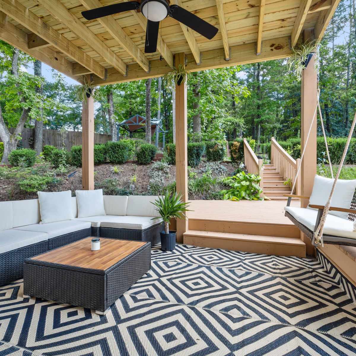 A covered outdoor seating area with a sectional sofa, a coffee table, and a swing, surrounded by greenery and leading to a wooden staircase.
